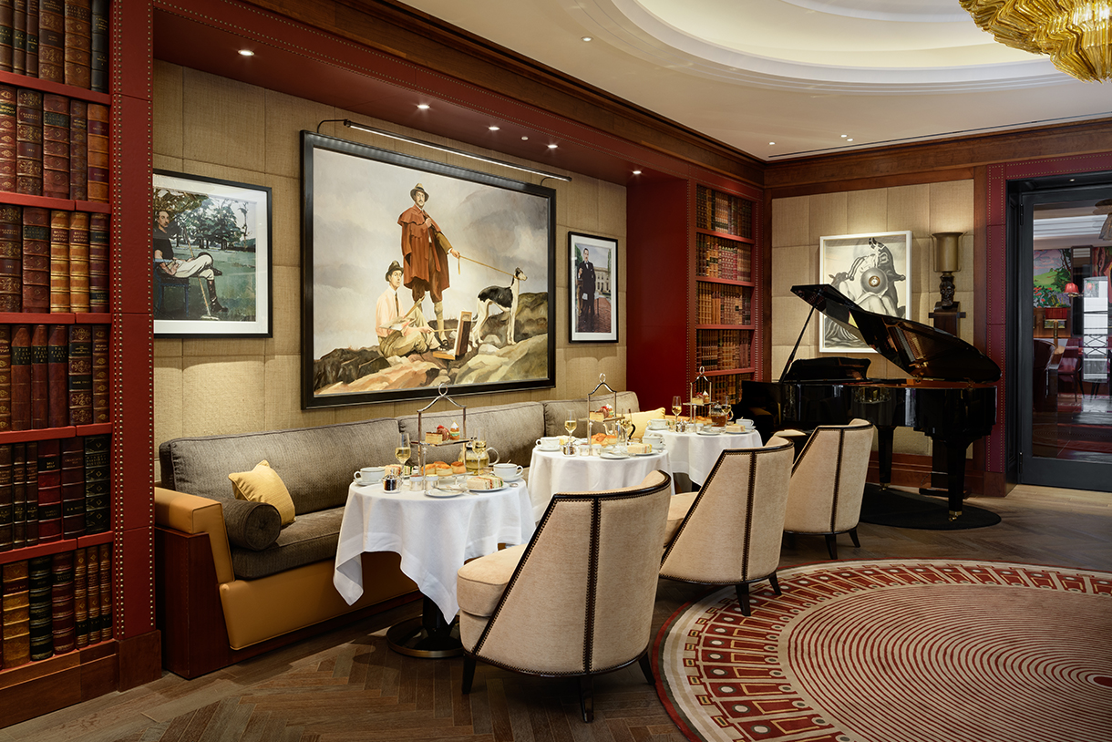 Gatsby tea room at The Beaumont with small dining tables, chairs and a long couch against the wall, paintings, bookshelves, and a grand piano.